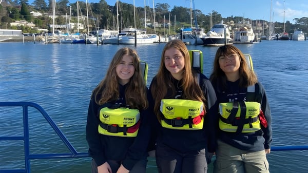 Tres estudiantes de la Le Fevre High School llevan chalecos salvavidas amarillos frente a un puerto con barcos.