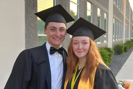 Dos graduados están frente a un edificio de la Leamington District Secondary School en togas de graduación y birretes.