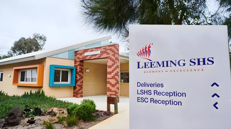El moderno edificio de una sola planta de la Leeming Senior High School con fachada de ladrillo y un cartel etiquetado está rodeado de árboles y arbustos.