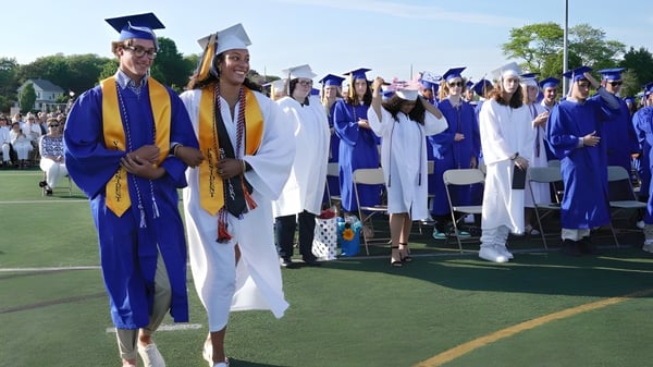 Un grupo de graduadas y graduados de la Leo Hayes High School está de pie en togas azules y blancas en un campo con árboles y edificios al fondo.