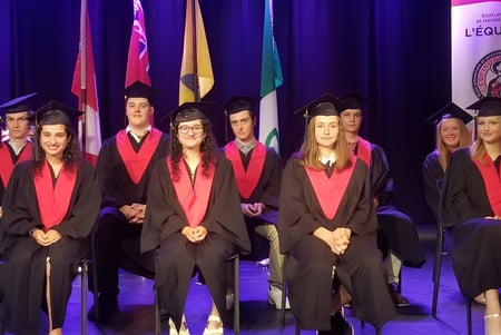 Las graduadas y graduados de la École élémentaire et secondaire publique L'Équinoxe están en vestimenta académica sobre un escenario en la ceremonia de graduación.