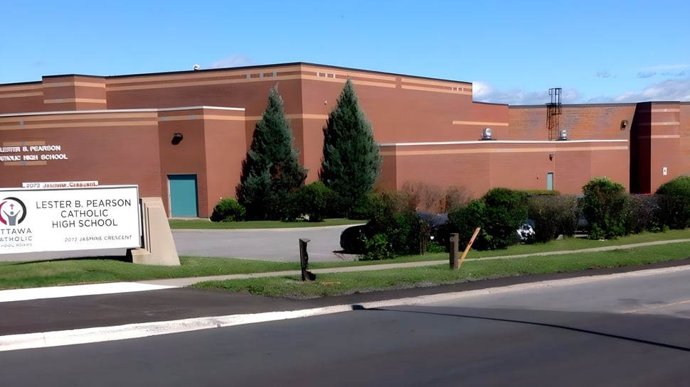 El gran edificio de ladrillo de la Lester B. Pearson Catholic High School con un letrero frente a árboles y un estacionamiento al fondo.