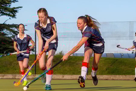 Las alumnas de Leweston School juegan un partido de hockey en un campo deportivo con bosque y agua de fondo.