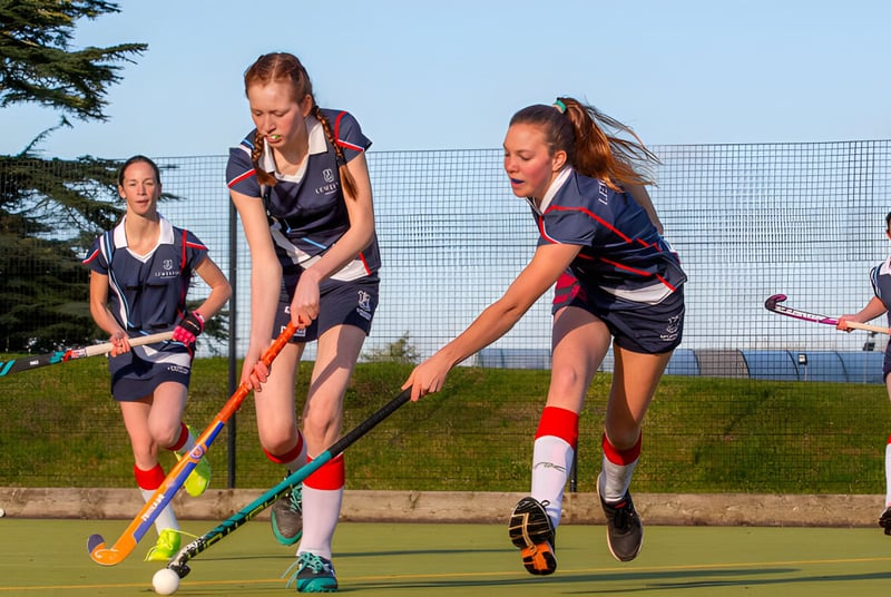Las alumnas de Leweston School juegan un partido de hockey en un campo deportivo con bosque y agua de fondo.