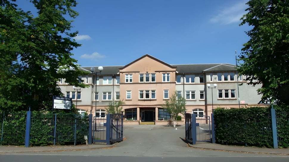 El edificio de ladrillo de varios pisos de la Lomond School con techo a dos aguas está rodeado de un entorno verde y un camino pavimentado con puertas en primer plano.