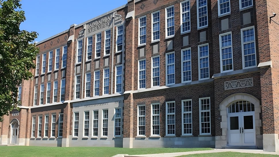El edificio de ladrillo de varios pisos con muchas ventanas y una entrada arqueada del London South Collegiate Institute está rodeado de árboles.