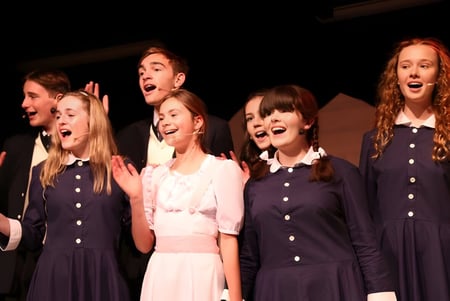 Un grupo de alumnas del Long Bay College actúa en el escenario con ropa oscura bajo una iluminación dramática.
