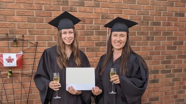 Dos estudiantes de la Longfields-Davidson Heights Secondary School posan con diplomas y copas de champán frente a una pared de ladrillo con una bandera canadiense.