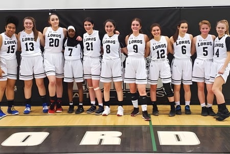 Un grupo de jóvenes jugadoras de baloncesto en camisetas blancas está en el campo de baloncesto de la Lord Beaverbrook High School.