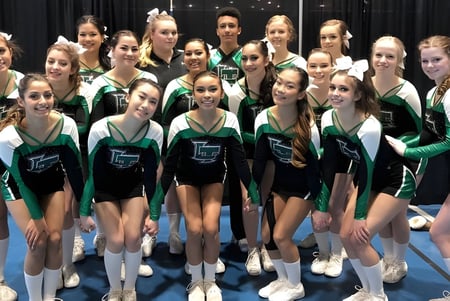 Un grupo de jóvenes gimnastas de la Lord Tweedsmuir Secondary School posan juntas en uniformes verdes y negros en un escenario.