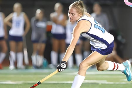 Una atleta de la Loreto High School Beaufort corre en un campo deportivo con otras atletas al fondo.