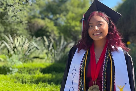 Una joven sonriente en birrete y toga de graduación está en el área verde del Los Angeles Unified School District.