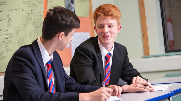 Dos estudiantes de la Loughborough Grammar School conversan en un escritorio en el aula con una pizarra al fondo.
