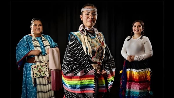 Tres estudiantes de Luther College High School visten ropa tradicional nativa americana con patrones coloridos y tocados.