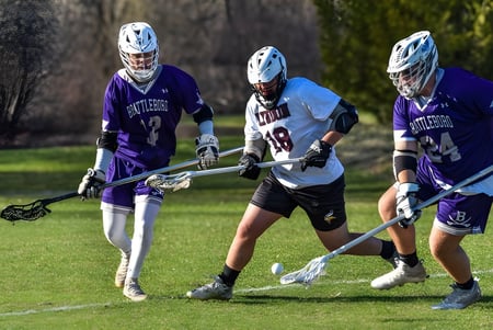 Tres jugadores en uniformes de lacrosse compiten en el campo deportivo del Lyndon Institute por la noche.