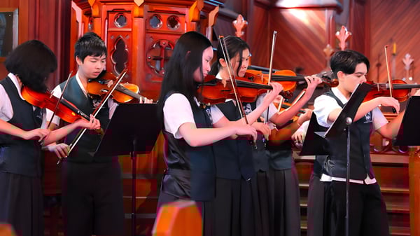 Un grupo de músicos en ropa formal actúa en el escenario del Lynfield College frente a un fondo rojo y dorado.