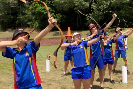Estudiantes de la Malanda State High School practican tiro con arco al aire libre en un campo deportivo rodeado de vegetación.