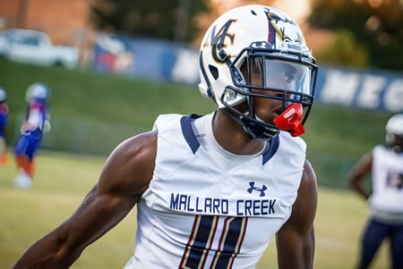 Un futbolista en uniforme blanco de la Mallard Creek High School está en el campo con otros jugadores y espectadores al fondo.