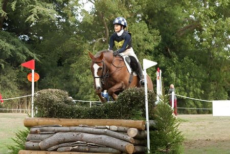 Una estudiante de la Malvern St. James Girls' School monta en una carrera de caballos sobre un obstáculo en un paisaje forestal cubierto de hierba.