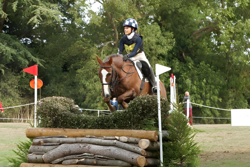 Una estudiante de la Malvern St. James Girls' School monta en una carrera de caballos sobre un obstáculo en un paisaje forestal cubierto de hierba.