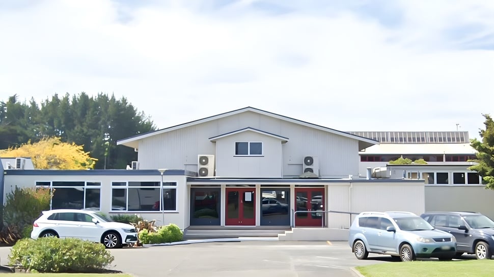 El moderno edificio blanco con techo inclinado en el campus del Manawatu College está rodeado de vehículos estacionados y árboles.