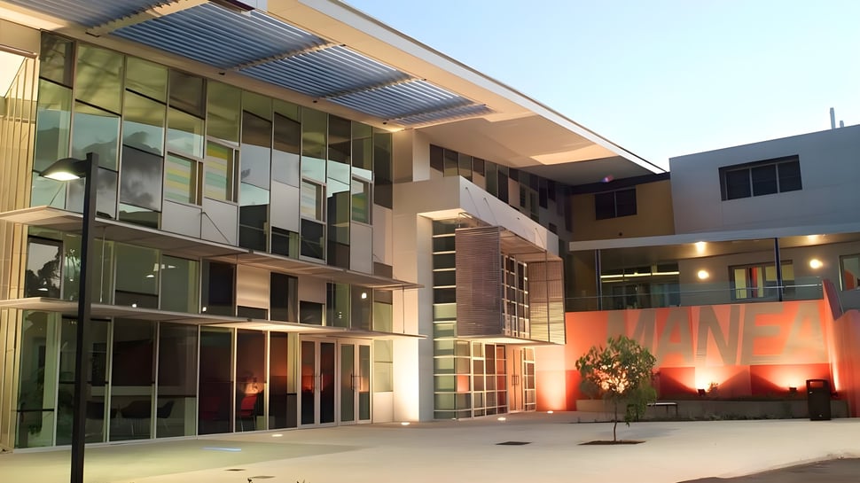 El moderno edificio del Manea Senior College muestra una fachada de vidrio con entrada cubierta y área exterior iluminada en un patio pavimentado.