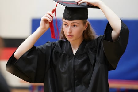 Una absolvente de la École secondaire publique Marc-Garneau está en la sala frente a un fondo azul con su manto y gorra de graduación negra en las manos.