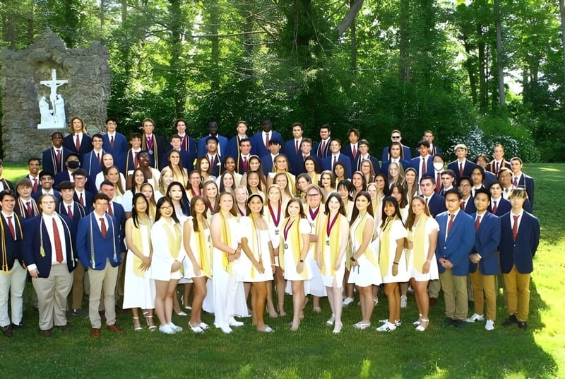 Un grupo de estudiantes de la Marianapolis Preparatory School está vestido con ropa formal frente a un monumento en forma de cruz al aire libre.