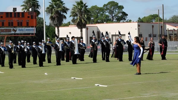 Un grupo de estudiantes uniformados de Marion County Public Schools está en formación en un campo de césped con palmeras y edificios al fondo.
