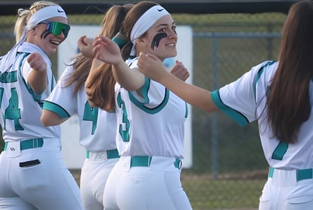 Un grupo de jóvenes atletas en uniformes blancos y verdes está en un campo de deporte celebrando juntas en el campus de Marion County Public Schools.