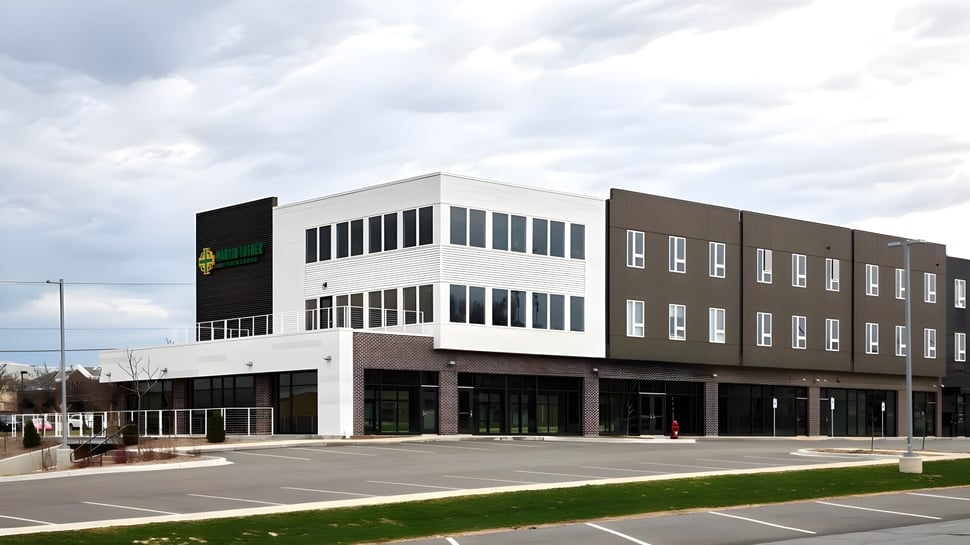 El moderno edificio de la Martin Luther High School con fachada de vidrio y ladrillo se encuentra en un estacionamiento pavimentado bajo un cielo nublado.