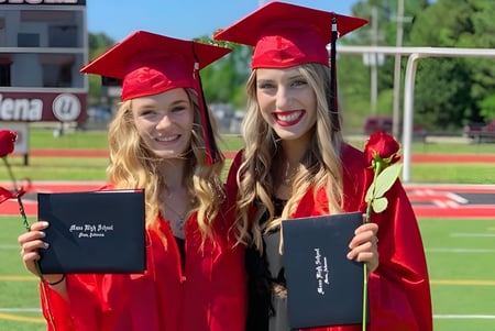 Dos graduadas de la Mena High School están en el campo deportivo sosteniendo sus diplomas.
