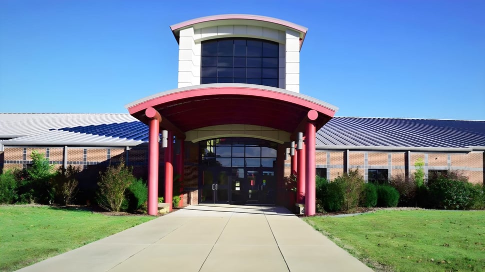 El moderno edificio de varios pisos de la Mena High School con entrada de vidrio y un toldo rojo se encuentra en un área de césped bajo un cielo despejado.