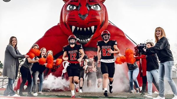 Atletas de la Mena High School caminan a través de nubes de humo bajo la gran figura del mascota roja.