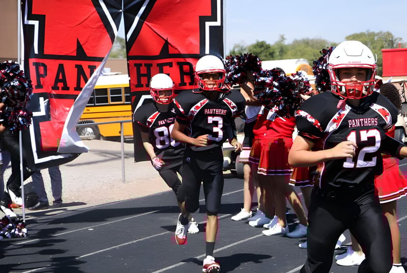 Un grupo de jugadores de fútbol de la Menaul School está frente a un gran banner de Panthers con un autobús escolar y espectadores al fondo.