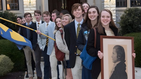 Un grupo de estudiantes de la Mercersburg Academy está con un retrato enmarcado frente a un edificio con una bandera.