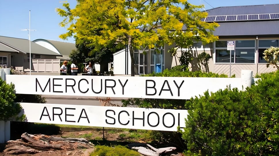 El cartel de la Mercury Bay Area School está frente a árboles con un edificio escolar al fondo.