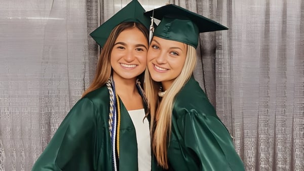 Dos estudiantes de la Mercyhurst Prep School en togas de graduación verdes posan frente a una cortina gris.