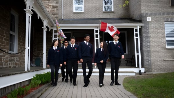 Estudiantes de la Merrick Preparatory School en ropa formal caminan juntos por un camino frente a un edificio de ladrillo con la bandera canadiense.