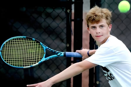 Un estudiante de la Merrick Preparatory School está con una raqueta de tenis frente a la cerca de alambre en el campo de tenis.