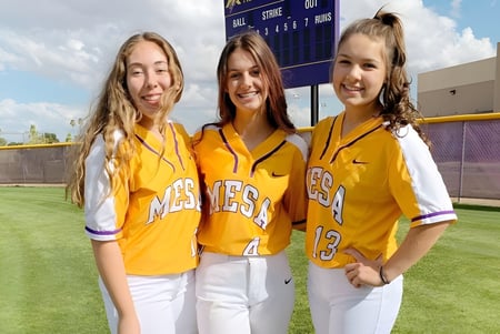 Tres estudiantes de la Mesa High School están en camisetas deportivas amarillas en un campo de béisbol con un marcador en el fondo.