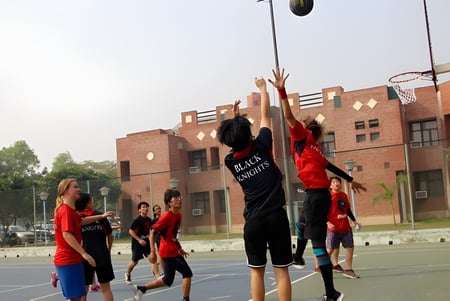 Estudiantes de la Metro International Secondary Academy juegan al baloncesto en una cancha exterior frente a edificios de ladrillo.