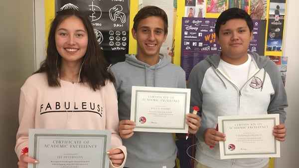 Tres estudiantes de la Miami Beach Senior High School sostienen certificados en un aula con carteles en las paredes.