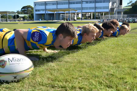 Un grupo de estudiantes de la Miami State High School hace flexiones en un campo deportivo frente a una gran instalación deportiva.