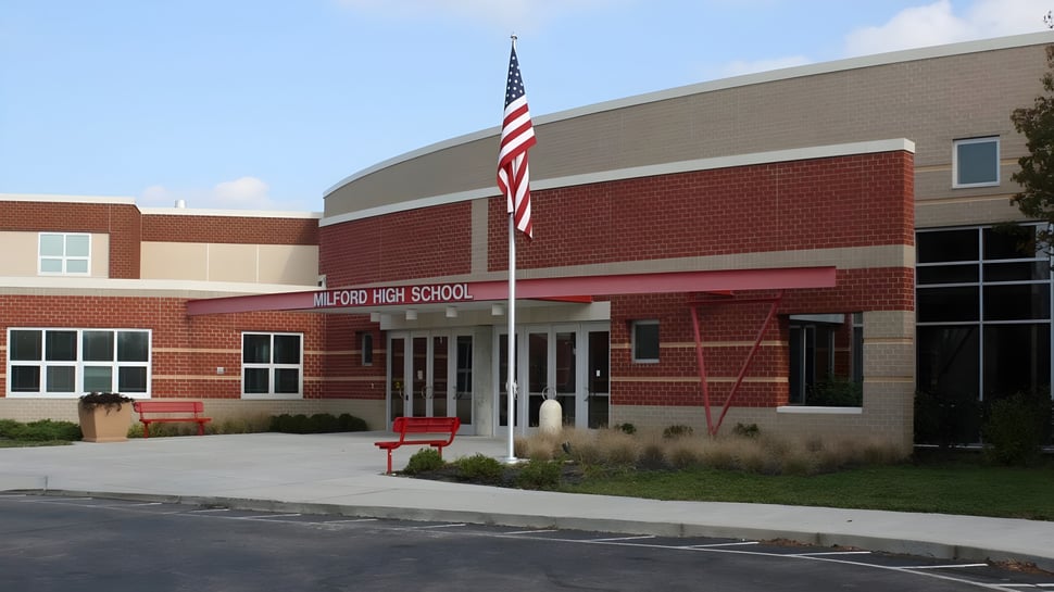 El moderno edificio de ladrillo y vidrio del Milford Public School District está rodeado de un estacionamiento y una bandera americana en el techo.