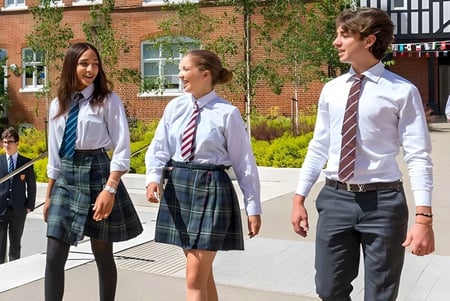 Tres estudiantes en uniformes escolares están frente a un edificio de ladrillo con paredes cubiertas de hiedra en el campus de la Mill Hill International.