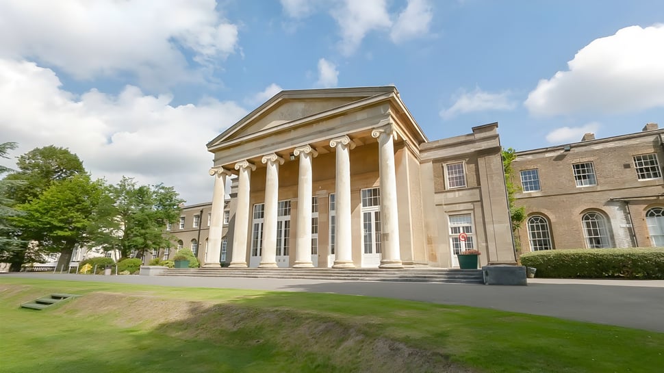 El clásico edificio escolar de la Mill Hill School con columnas y un césped en primer plano bajo un cielo nublado.