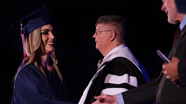 Una joven graduada de la Millfield School le da la mano a un hombre mayor en toga académica durante una celebración.