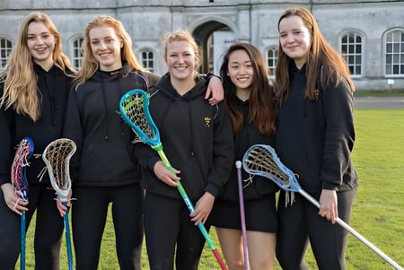 Un grupo de alumnas de la Milton Abbey School está con palas de lacrosse frente a un edificio histórico con ventanas de arco.