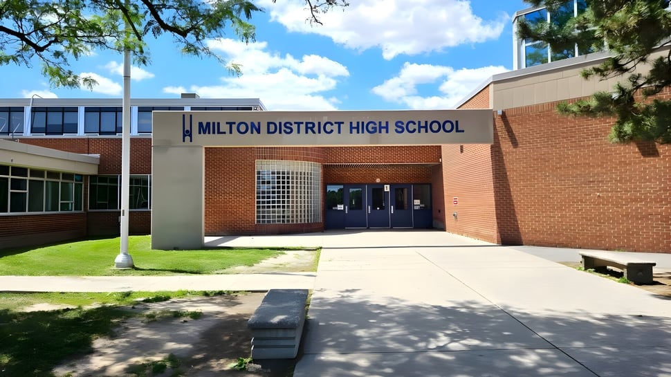 El edificio de ladrillo de la Milton District High School está rodeado de áreas de césped y árboles bajo un cielo azul con nubes.